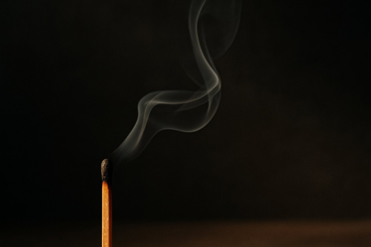 Fiammifero appena spento ripreso da vicino, con un filo di fumo che sale in luce laterale calda su fondo scuro