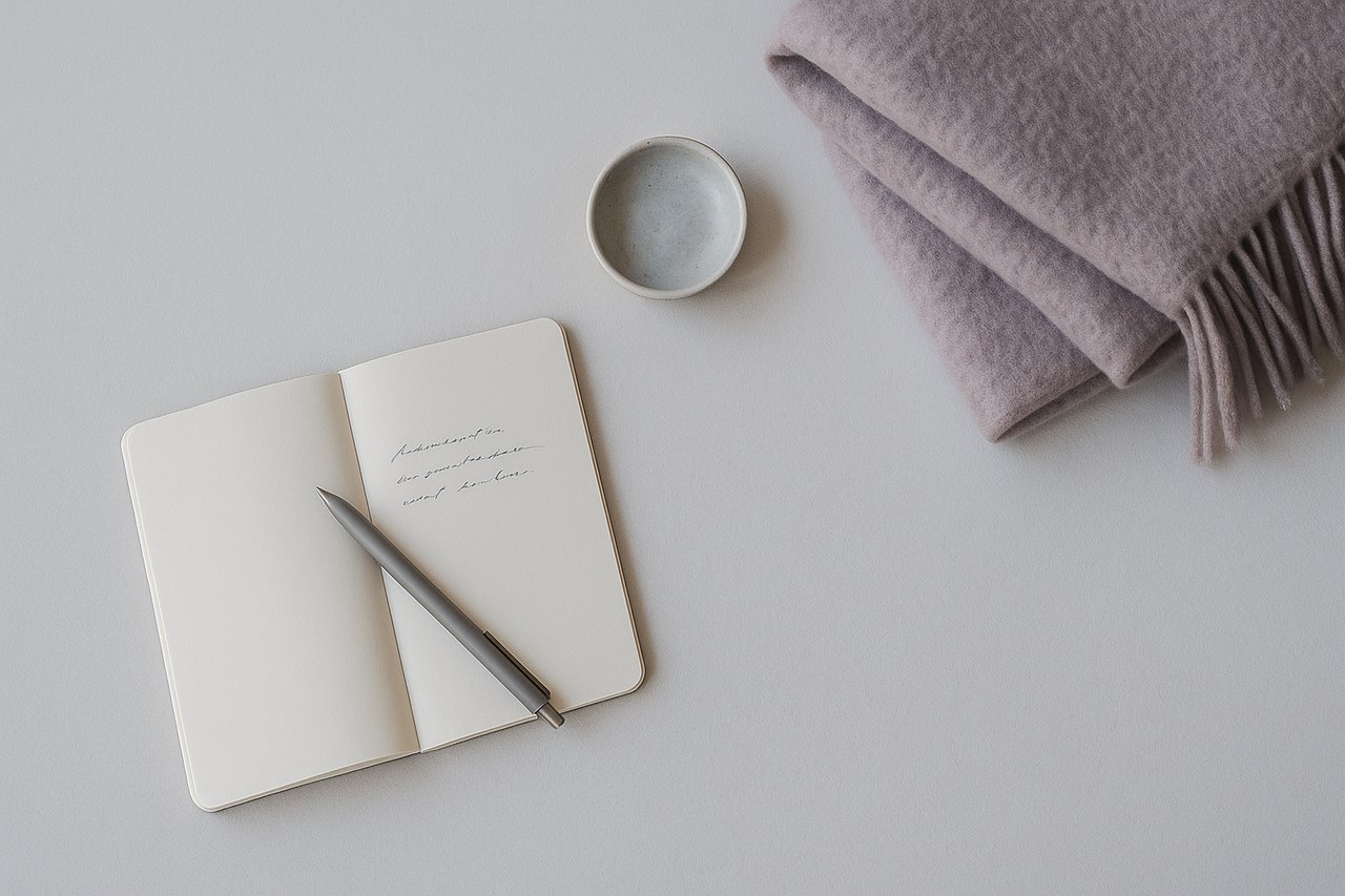 Quaderno aperto con una penna appoggiata, una coperta di lana ripiegata e una piccola ciotola di ceramica, ripresi dall'alto in luce fredda diffusa